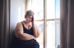 Eine depressive, einsame, korpulente Frau in Unterwäsche sitzt am Fenster, schaut nach unten und denkt nach, zu Hause. Bild zum Thema Kraft und Energie tanken