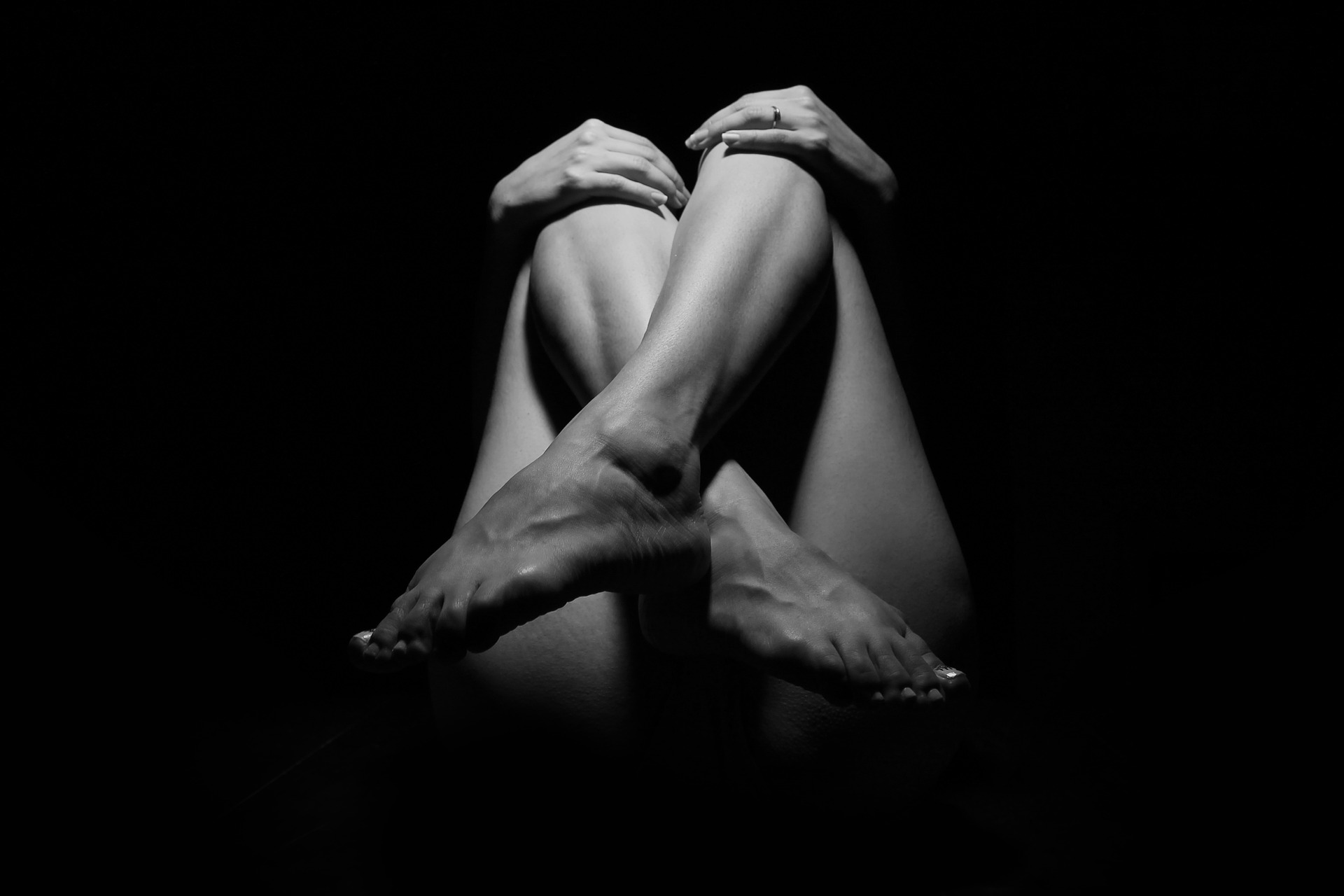 Schwarz-weißer Akt einer Frau mit gekreuzten Beinen, die ihre Knie hält Genitalien im Dunkeln betrachten. Bild zum Thema: Allround-Talent Eigenfett!